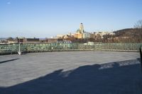 some benches are empty on the concrete patio near a fence and large city skyline in the background