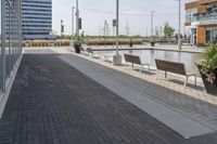 the empty brick path has several plants and two benches near by it with the building behind them