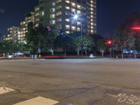 an empty city street lit up at night with traffic in the foreground, and many buildings, trees, and bushes