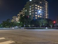 an empty city street lit up at night with traffic in the foreground, and many buildings, trees, and bushes