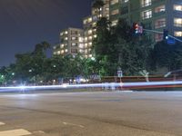 an empty city street lit up at night with traffic in the foreground, and many buildings, trees, and bushes