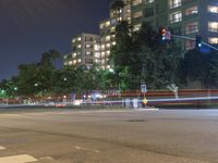 an empty city street lit up at night with traffic in the foreground, and many buildings, trees, and bushes