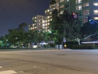 an empty city street lit up at night with traffic in the foreground, and many buildings, trees, and bushes