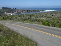 there is an empty paved road by the ocean with grass and bushes in the foreground