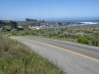 there is an empty paved road by the ocean with grass and bushes in the foreground