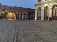 an empty cobblestone patio with a parking lot and building in the background with a few lights