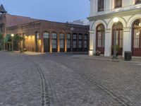 an empty cobblestone patio with a parking lot and building in the background with a few lights