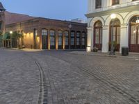 an empty cobblestone patio with a parking lot and building in the background with a few lights