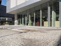 an empty brick road in front of a modern building with large glass windows and doors