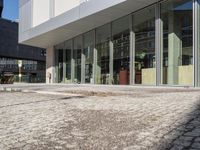 an empty brick road in front of a modern building with large glass windows and doors