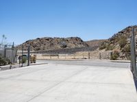 empty concrete area with fence and barbed wire fencing near wall and mountains on hill with blue sky in background