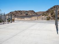 empty concrete area with fence and barbed wire fencing near wall and mountains on hill with blue sky in background