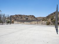 empty concrete area with fence and barbed wire fencing near wall and mountains on hill with blue sky in background