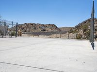 empty concrete area with fence and barbed wire fencing near wall and mountains on hill with blue sky in background