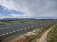 an empty country road in the middle of nowhere's countryside under cloudy skies with mountains behind