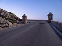 an empty country road with a clock on the gate to the hill side and rocks in the background