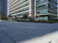 an empty courtyard in front of a very tall glass building with trees, plants and grass