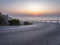 an empty curved road next to the water with a sunset behind it on the sea