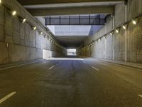 an empty dark street lined with concrete walls and lights above it in a tunnel with the road going under