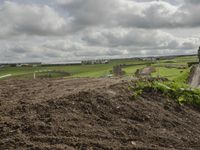 an empty dirt pile in a field near a road and farm area that is in the distance with a green field, trees and buildings