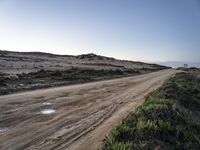 an empty dirt road on a beach covered in bushes and sand with water puddles