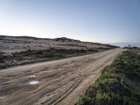 an empty dirt road on a beach covered in bushes and sand with water puddles