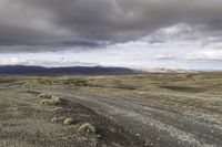 an empty dirt road running in the desert with mountains in the background, on a cloudy day