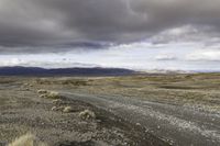 an empty dirt road running in the desert with mountains in the background, on a cloudy day