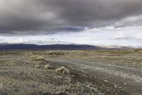 an empty dirt road running in the desert with mountains in the background, on a cloudy day