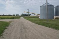 an empty dirt road and silos in the background with corn on either side, and a yellow object in the distance