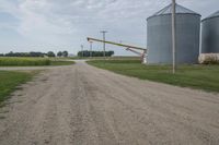 an empty dirt road and silos in the background with corn on either side, and a yellow object in the distance