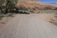 an empty dirt road in the desert with a house and trees by it with no people