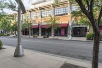 an empty downtown street with lots of buildings and trees in the foregrounds and a sidewalk next to some shops on the other side