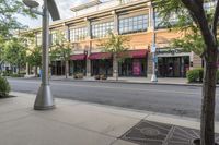 an empty downtown street with lots of buildings and trees in the foregrounds and a sidewalk next to some shops on the other side