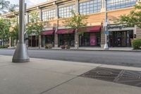 an empty downtown street with lots of buildings and trees in the foregrounds and a sidewalk next to some shops on the other side