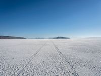 the tracks of a person walking across a large empty field of snow and mountains in the distance