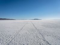 the tracks of a person walking across a large empty field of snow and mountains in the distance