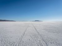 the tracks of a person walking across a large empty field of snow and mountains in the distance