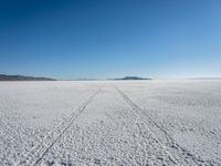 the tracks of a person walking across a large empty field of snow and mountains in the distance