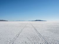 the tracks of a person walking across a large empty field of snow and mountains in the distance