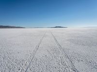 the tracks of a person walking across a large empty field of snow and mountains in the distance