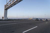 several cars drive on an empty freeway under a suspension bridge in the morning sun setting