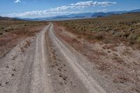 an empty gravel road with mountains in the distance and grass on either side of the road