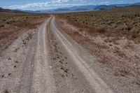 an empty gravel road with mountains in the distance and grass on either side of the road