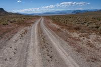 an empty gravel road with mountains in the distance and grass on either side of the road