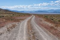 an empty gravel road with mountains in the distance and grass on either side of the road