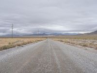 a gravel road is empty in the desert near a mountain range and power lines, with telephone poles in front