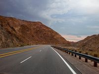 view of an empty highway going through a desert on a cloudy day with clouds and hills