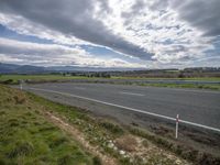 a sky filled with some clouds next to a empty highway and green hills with some bushes