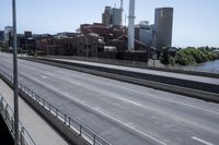 an empty highway running along side a city with tall buildings in the background and other large building, including a large factory, and an old train bridge